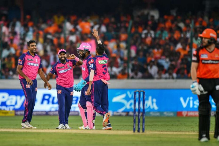 Cricket players in pink uniforms celebrate on the field as a crowd cheers in the stands behind them, with a batsman in orange nearby.
