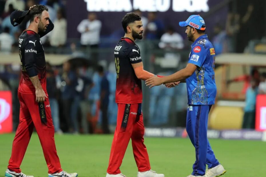RCB vs MI - Two cricket players from opposing teams shake hands on the field after a match; one in blue, the other in red and black, with blurred spectators in the background.